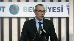 Takım elbiseli uzun seyrek saçlı bir erkeke mikrofona konuşuyor. Arkada belediyesi ifadesi yazıyor.