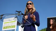 Annie Farmer, con un micrófono en la mano, habla en un atril. Lleva gafas de sol y viste de azul. Hace sol y el atril muestra el mensaje "Junto a las víctimas" escrito en inglés.