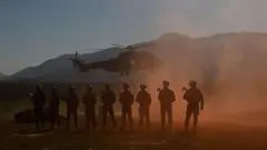 A imagem mostra a silhueta de oito militares armados alinhados em primeiro plano enquanto um helicóptero militar paira ao fundo, levantando poeira. Ao fundo, há montanhas sob a luz do entardecer, compondo um cenário de treinamento ou operação militar.