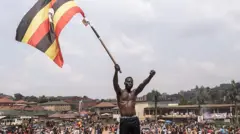 Wani matashi marar riga ya tsaya a saman wani ƙaramin tudu, yana ɗaga tutar Uganda. A bayansa akwai dandazon jama'a a wani gangamin da ɗan adawa Robert Kyagulanyi Ssentamu ya jagoranta.