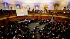 Di Archbishop of Canterbury address di General Synod. She stand behind a long table with her image on a large screen above her. Plenti pipo dey in attendance. 