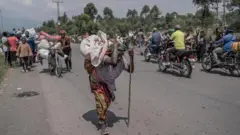 Umugore wikoreye bimwe mu byo atunze, mu gihe abantu bari barimo guhunga bava muri teritwari ya Masisi, mu ntara ya Kivu ya Ruguru, nyuma y'imirwano ikaze hagati y'inyeshyamba za M23 n'ingabo za Repubulika ya Demokarasi ya Congo, aha ni mu muhanda uri hafi y'i Sake, ku itariki ya 7 Gashyantare (2) mu mwaka wa 2024. 