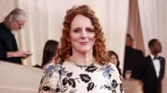 Maggie O'Farrell tem cabelos ruivos e cacheados, meio presos, e veste um vestido branco com estampa de flores em preto, branco e azul. Ela sorri para a câmera enquanto posa no tapete vermelho