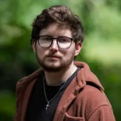 Homem de casaco marrom com camiseta preta e um crucifixo, em pé, usando óculos em frente a um arbusto