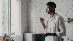 Homem se prepara para tomar um comprimido, segurando um copo d'água e olhando para a janela