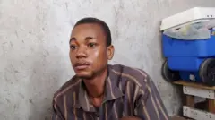 Wilson, un joven negro de pelo corto y negro, con una camisa marrón a rayas, mira a la izquierda de la cámara con expresión pensativa. A su izquierda hay un contenedor de plástico azul con ruedas, apilado sobre un mueble de madera.