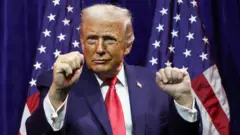 Президент Трамп сжимает руки в кулаки и держит их на высоте плеч, выступая перед однопартийцами. На заднем плане два американских флага