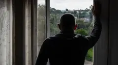 مردی از پنجره به بیرون نگاه می‌کند، دست راست بالای پنجره تکیه داده و خانه‌ها، درختان و فضای سبز در بیرون. 