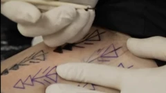 Pele nua com setas tatuadas em azul e preto, algumas preenchidas. Duas mãos com luvas de látex estão sobre a pele. Em uma das mãos, uma agulha de tatuagem está posicionada para uso.