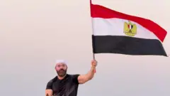 المدون المصري، رامي عادل يمتطي حصاناً ويرفع بيده اليسرى علم مصر، وحوله رمال وأشجار.