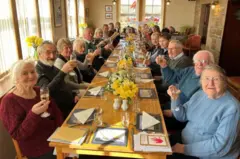 Longa mesa com familiares sentados, erguendo taças de champanhe