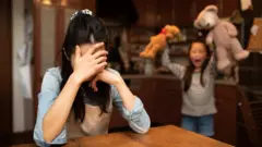 Seorang perempuan yang menyandarkan kepalanya dengan tangan dan seorang akan memegang boneka.