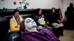 Mulher reclinada com toalha na cabeça e creme para clarear a pele no rosto, com outras mulheres desfocadas ao fundo