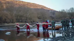 長袖と長ズボンの赤い作業着を着た男性たちが、白くて長いオイルフェンスを担いで川の中へ入っている