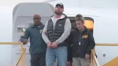 Ryan Wedding es escoltado por dos agentes del FBI mientras desciende de un avión