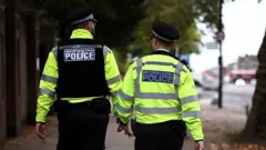 Двое сотрудников Лондонской полиции патрулируют улицы в Стэнфорд-Хилл на севере Лондона 2 октября 2025 года. Они повернуты к камере спиной и одеты в униформу и светоотражающие жилеты.