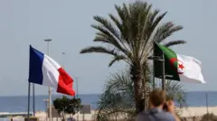 Drapeaux nationaux de la France et de l'Algérie flottant en Algérie, avec un palmier au milieu et l'océan en arrière-plan.