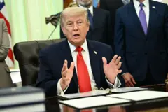الرئيس الأمريكي، دونالد ترامب، مرتديا بدلة زرقاء وقميصا أبيض ورابطة عنق حمراء، ويتحدث وهو جالس داخل المكتب البيضاوي في البيت الأبيض، وخلفه عدد من المسؤولين الأمريكيين.