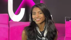 一頭長長的黑髮的安雅·薩拉·拉哈迪 (Anja-Sara Lahady) 面帶微笑，站在粉紅色墊子前。