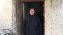 Siti Aminah na porta de sua casa, usando um hijab e um vestido preto.