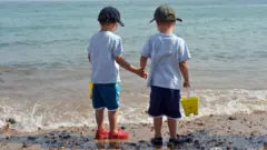 Duas crianças estão de costas, em uma praia. Elas carregam baldes e estão perto do mar.