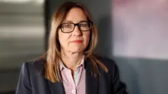 Rachel Horman-Brown, de óculos e usando um casaco escuro e camisa clara de botões, com fundo interno escuro, olhando para a câmera