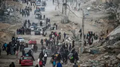 Pessoas caminham carregando sacolas e outros pertences em meio a escombros na Faixa de Gaza