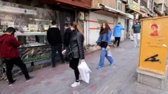 چند زن و مرد در حال عبور از مقابل یک مغازه ساعت‌فروشی با تصویری از علی خامنه‌ای، رهبر کشته‌شده ایران، روی یک ورق فلزی زرد رنگ در میدان تجریش تهران.
