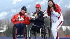 3選手が表彰台の上で横に並んでいる。全員笑顔で、左手でメダルを持ち、胸の前で見せている。背後には山並みが見える