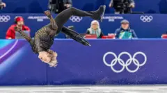 Ilia Malinin does a backflip on ice skates with judges and the Olympic logo in the background during an event.