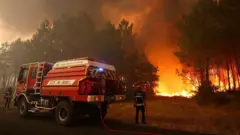 Fransa'da alevler içinde yanan bir ormanı söndürmek için yol kenarında durmuş bir itfaiye aracı ve ellerinde hortumlarla iki itfaiye görevlisi