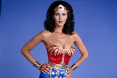 Lynda Carter con su característico disfraz de Wonder Woman. El atuendo tiene un top rojo y dorado, con pantalones cortos azules con estrellas blancas. Tiene una corona triangular dorada en el frente con una estrella roja, puños plateados con estrellas rojas y una cuerda dorada llamada lazo de la verdad que sale de su cinturón.
