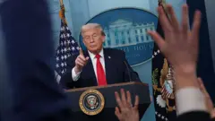 Donald Trump, soru sormak için el kaldıran gazetecilerin ellerinin arasından Beyaz Saray'ın kürsüsünde görülüyor. Başparmağını söz vereceği gazeteye uzatmak üzere kaldırmış