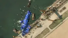 Imagen aérea por satélite de un barco en el puerto cubierto de tela azul, rodeado de embarcaciones más pequeñas.