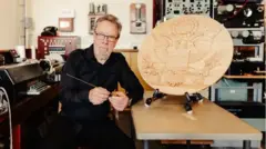 John Little é um homem mais velho, de cabelos grisalhos e óculos de grau. Ele está ao lado de um Grande Selo em madeira e segurando a réplica do dispositivo