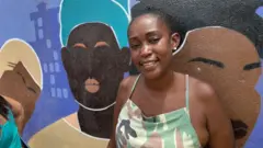 Taís Motta sorrindo em frente a muro grafitado com desenhos de mulheres pretas