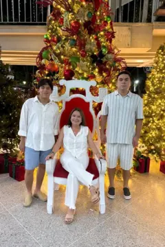 María Lourdes Pérez, vestida de branco e de óculos, sentada em uma cadeira vermelha e branca, com seus filhos Gonzalo à direita e Santiago à esquerda, em frente a três árvores de Natal decoradas e iluminadas