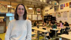 Una mujer morena con el pelo corto sonríe a la cámara frente a un aula llena de adolescentes. Está sonriendo y lleva un jersey gris. Hay alrededor de una docena de niños en la clase.