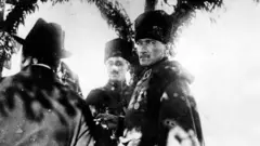 Mustafa Kemal, silah arkadaşlarıyla birlikte yandan görünüyor fakat ekrana bakarken çekilmiş, siyah beyaz, tarihsiz. 