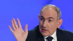 Ermenistan Başbakanı Nikol Paşinyan elini kaldırmış, arkada mavi bir fon