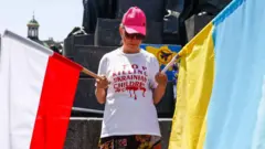 Жінка у білій футболці з червоними написами, у кепці та в окулярах, тримає два прапори - України та Польщі 