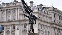 Скульптура: мужчина в костюме шагает вперед с постамента, одной рукой держа флаг, и этот же флаг закрывает его лицо. 