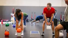Mujer joven levantando una pesa rusa de color naranja mientras entrena con una amigo en el gimnasio