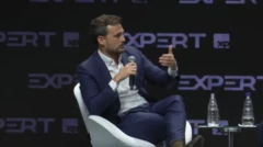 O economista João Landau, sócio-fundador da Vista Capital, durante sua fala no evento Expert XP, em 24 de julho de 2026. Ele veste terno azul sem gravata e camisa branca, e está sentado em uma cadeira branca, enquanto gesticula e segura um microfone. Ao lado dele, há uma pequena mesa de vidro, com duas garras plásticas de água em cima