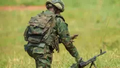 Umusirikare w'u Rwanda upfukamishije ivi rimwe mu byatsi, ukuboko kumwe gufashe ku mbunda irambitse hasi. Yambaye imyenda ya gisirikare irimo amabara y'icyatsi kibisi, khaki n'ikigina. Anambaye ingofero ya 'casque' ya gisirikare ndetse ahetse igikapu mu bitugu gisa n'imyenda ye. 