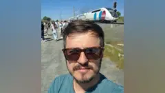 Jonathan Emery, vestindo uma camiseta e óculos de sol, está em frente a um trem que parou no meio da viagem na Espanha