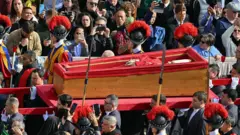Papa Francesco'nun cenazesi Aziz Pietro Meydanı'ndan geçiriliyor. Üzeri açık olan tabutu halk telefonlarıyla görüntülemeye çalışıyor. Tören kıyafetli askerler de tabuta eşlik ediyor