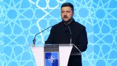 水色の背景の前、NATOの標章がついた演台に向かって話す、黒衣のゼレンスキー氏