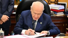 MHP Genel Başkanı Devlet Bahçeli, önündeki kağıtlara imza atıyor. Arkada ellerini önden bağlamış bir kişi görülüyor.