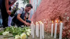 Homem branco acende vela em frente a um muro de escola, há rosas no chão